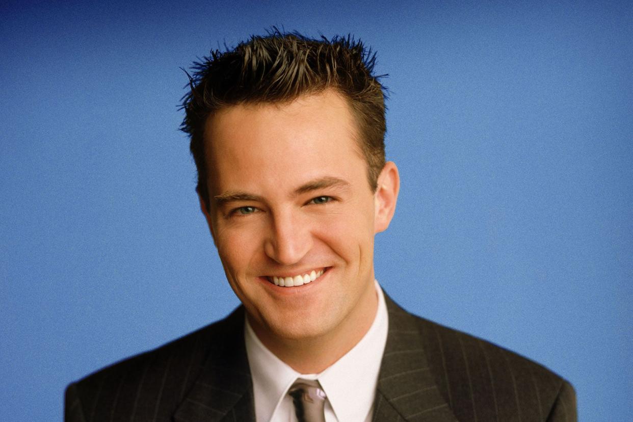 A twentysomething white man with spiked-up brown hair wearing a suit and tie against a blue background.