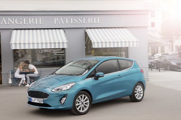 A teal green 2018 Ford Fiesta hatchback is shown parked in front of a French bakery