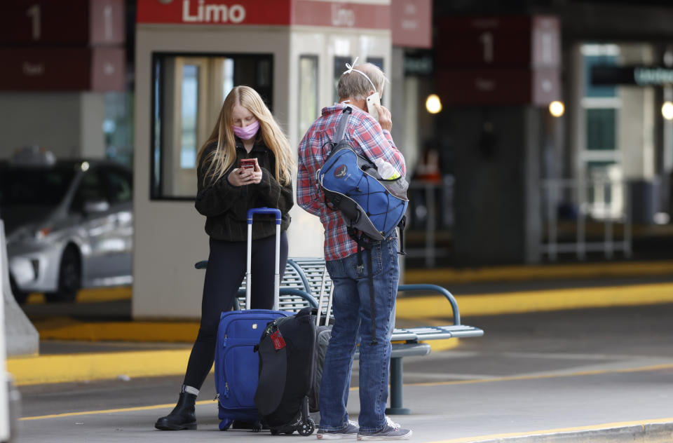 Travelers in face masks ait for a shuttle bus outside the main terminal after arriving at Denver International Airport Thursday, April 23, 2020, in Denver. (AP Photo/David Zalubowski)