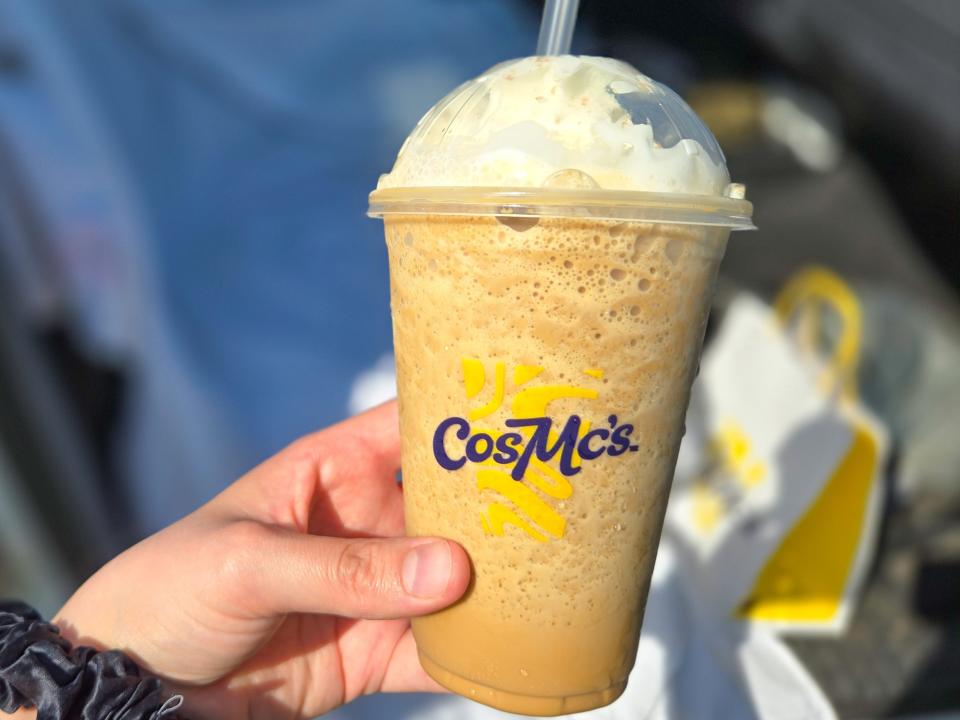 A hand holds a clear plastic cup with CosMc's logo and a dome cap with whipped cream in it. The cup is filled with a light-brown coffee