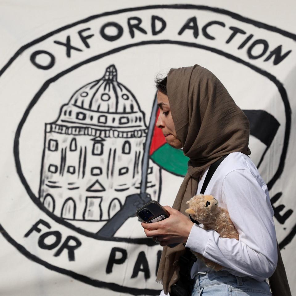 A young woman in a headscarf carries a soft toy past a banner reading 'Oxford Action for Palestine'