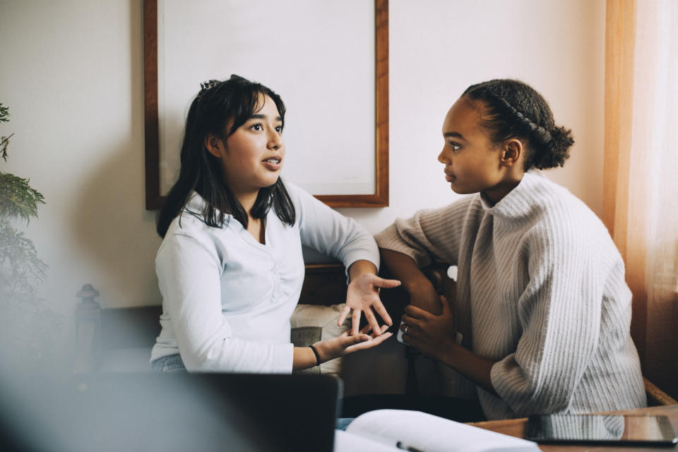 Two teenage girls sit on a sofa having a conversation, the South Asian girl on the left speaks and gestures with her hands while the Black girl on the right listens intently