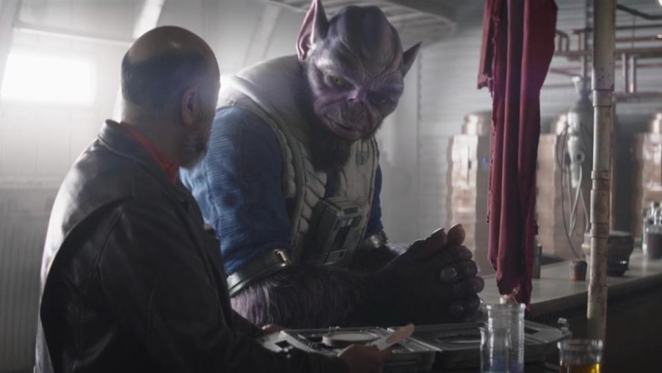 The large, big eared purple Lasat Zeb in his flight suit at a bar speaks with Carson Teva on The Mandalorian