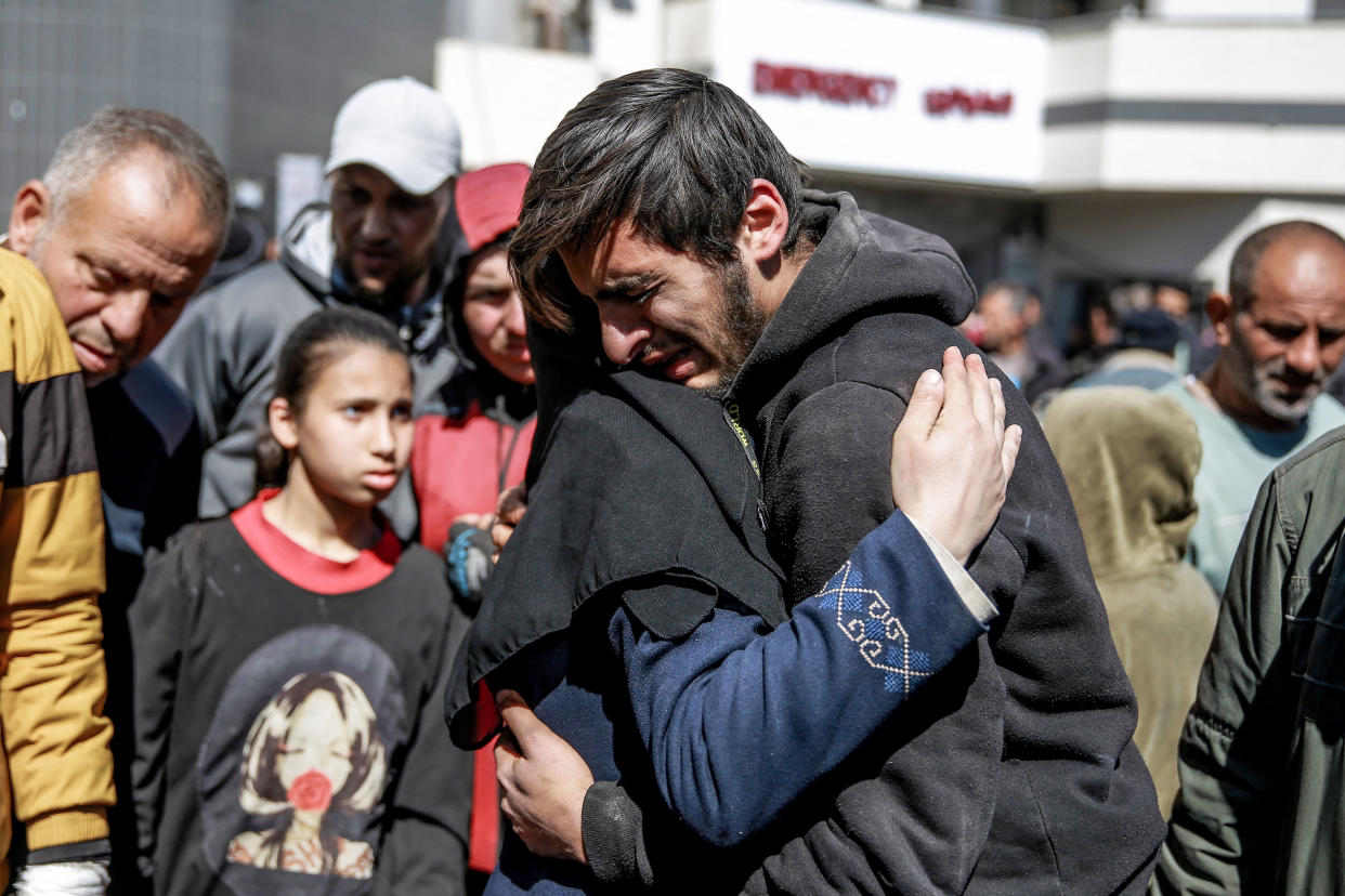 Two adults hug as they and several others mourn outside a hospital.