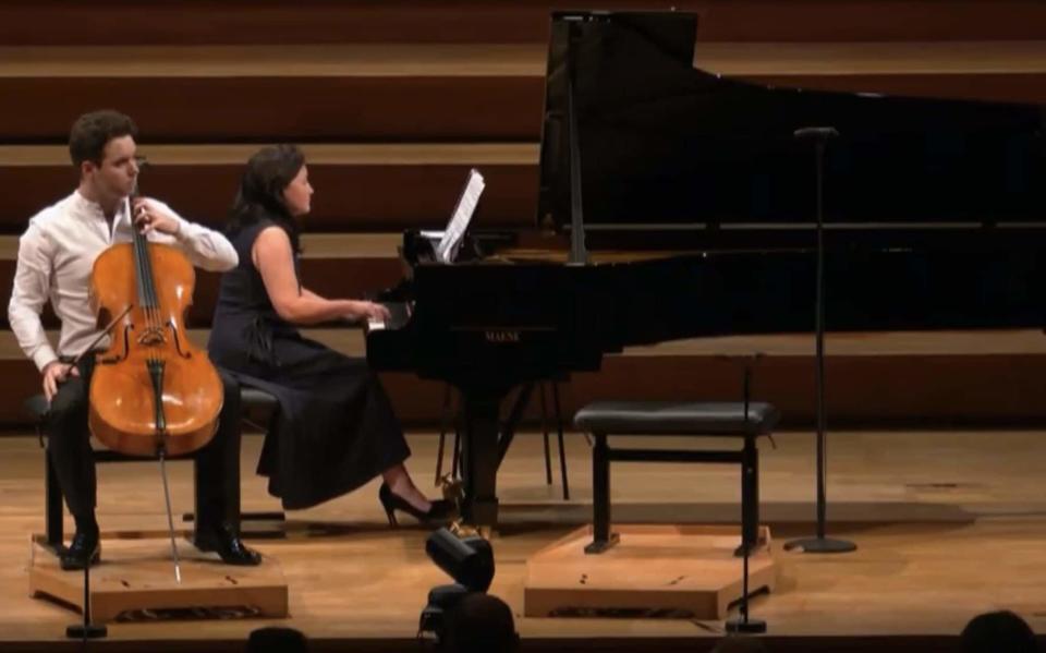 The young cellist sits on a stage with the instrument between his legs, next to a pianist