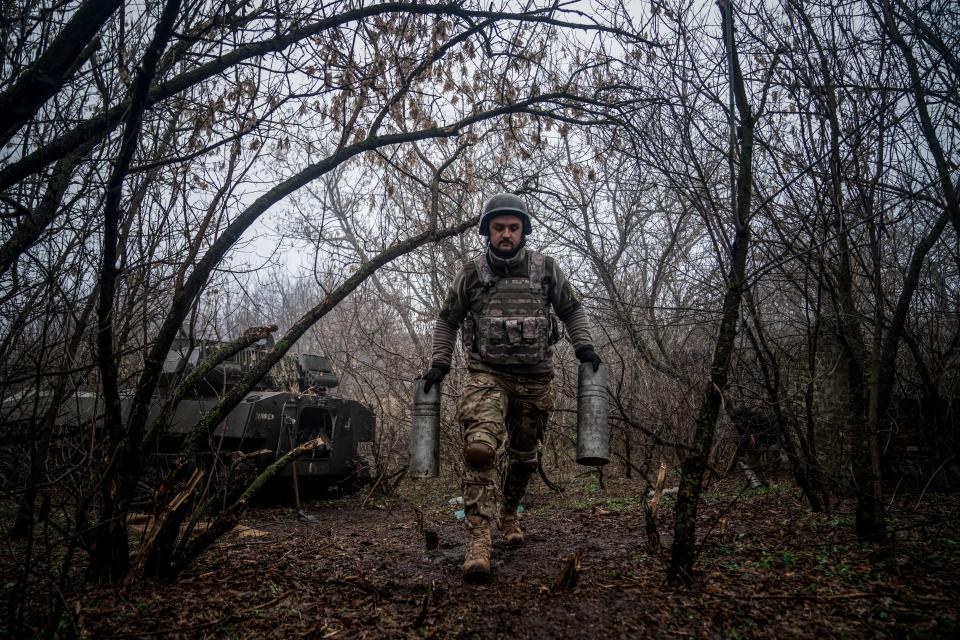 A Ukrainian artilleryman carries two empty artillery cartridge cases through the woods.