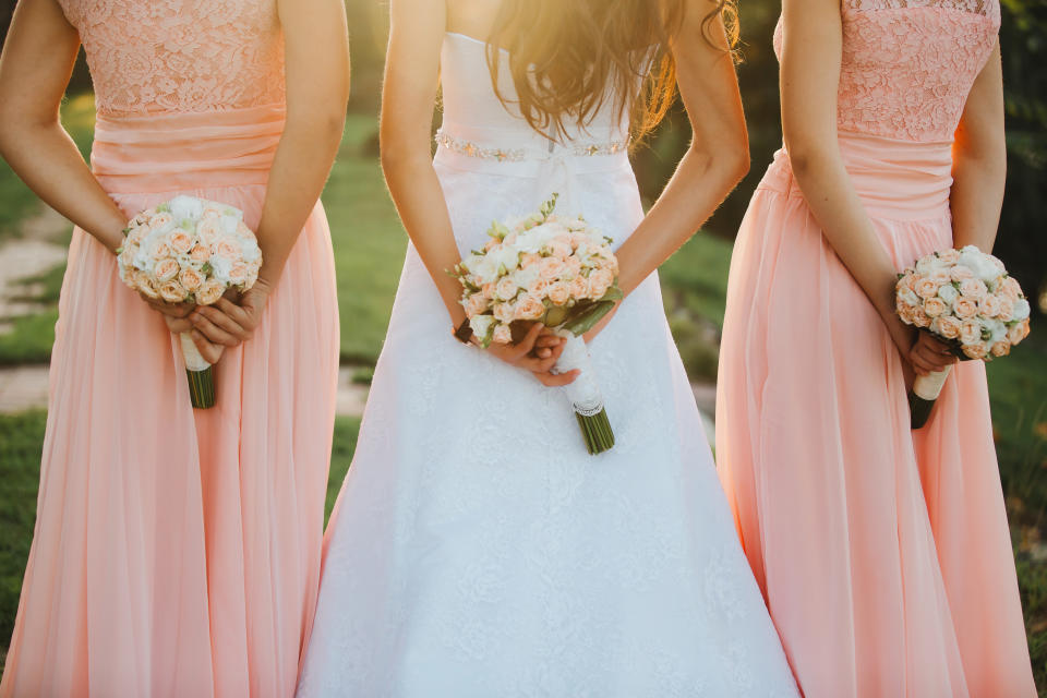 The bride and bridesmaids in an elegant dress is standing and holding hand bouquets of pastel pink flowers and greens with ribbon at nature. Young beautiful girls holds a wedding bouquet outdoors.