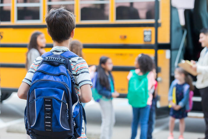 A child is seen from behind wearing a backpack and approaching a group of children near a school bus.