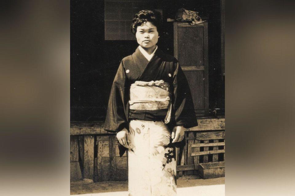 Black and white full length photo of Kiyomi Iguro. She has short dark hair and is wearing a traditional Japanese dress, of a black long sleeved top, and a white patterned wrap dress that stops at her feet. 