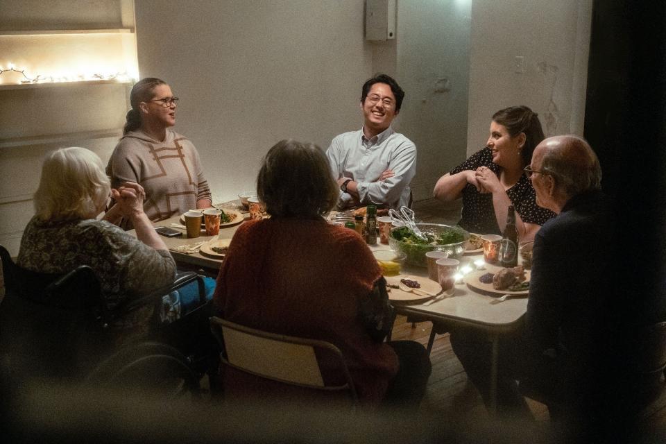 A white family, two adult sisters, one with an Asian boyfriend, their father, mother, and grandmother sit around a Thanksgiving table. Some laugh, some smile.