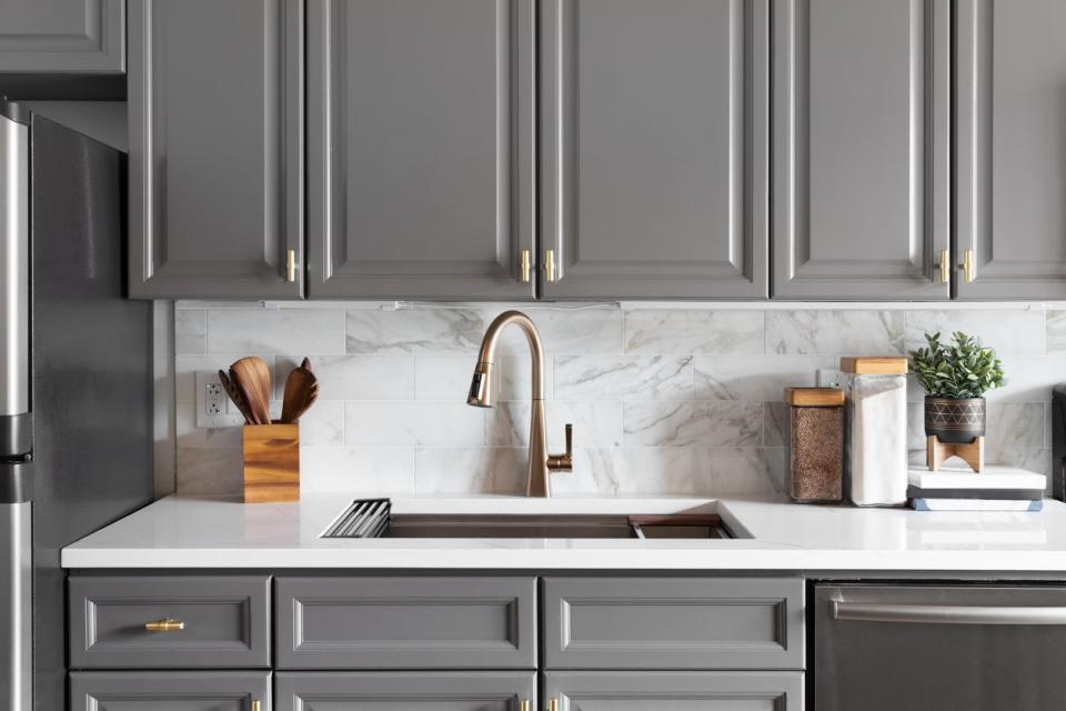 Grey kitchen cabinets with white countertop and brass sink faucet. 