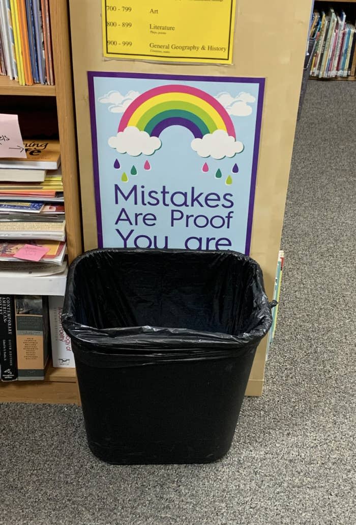 Sign reading "Mistakes Are Proof You are Trying" above a trash bin in a library by bookshelves