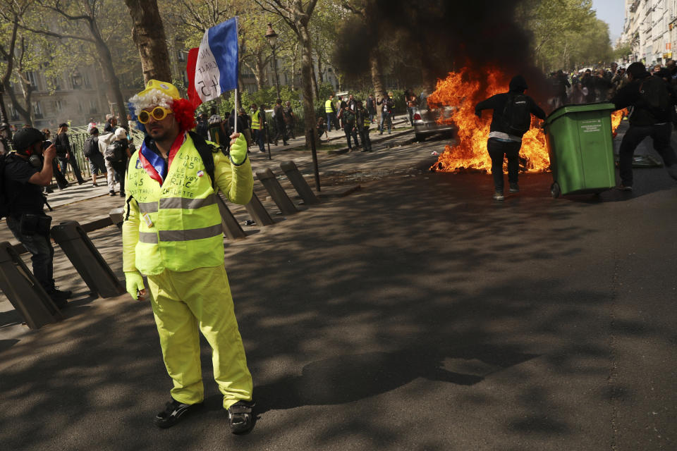 A protestors waves a French flag as a dumpster burns in the background during a yellow vest demonstration in Paris, Saturday, April 20, 2019. French yellow vest protesters are marching anew to remind the government that rebuilding the fire-ravaged Notre Dame Cathedral isn't the only problem the nation needs to solve. (AP Photo/Francisco Seco)