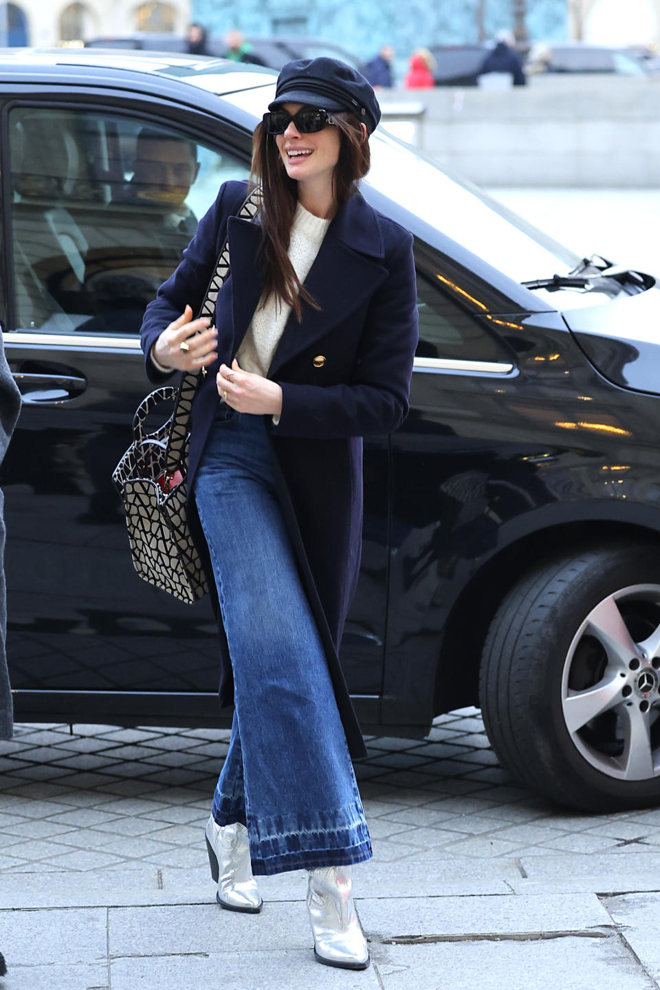 Anne Hathaway in a navy coat, white top, flared jeans, and silver boots steps out of a car