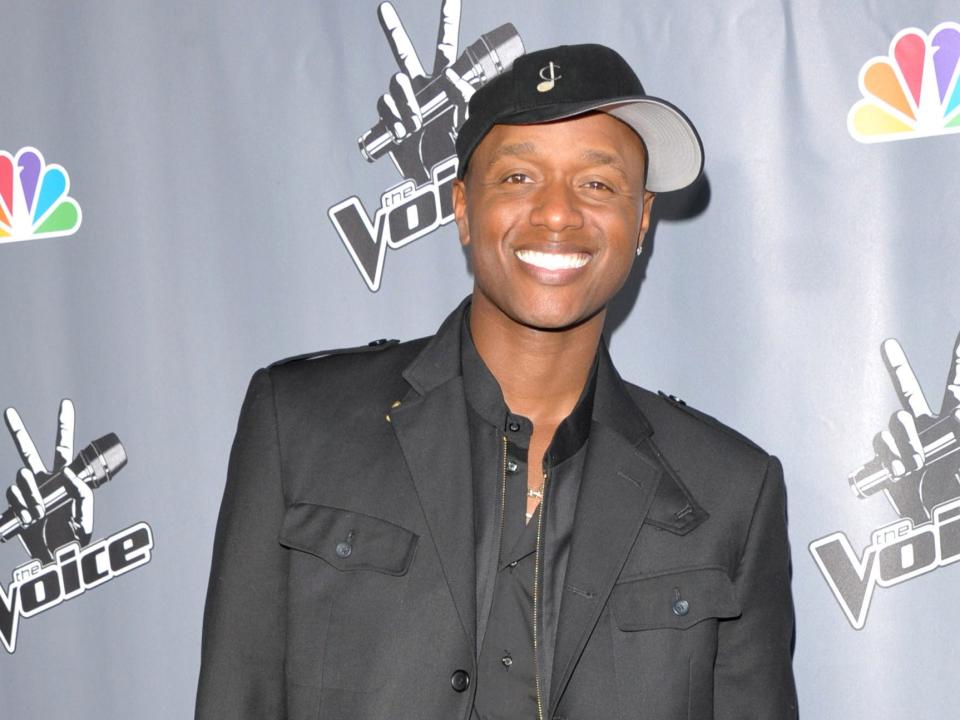 Javier smiling in a baseball cap, black shirt, and black jacket in front of a backdrop.