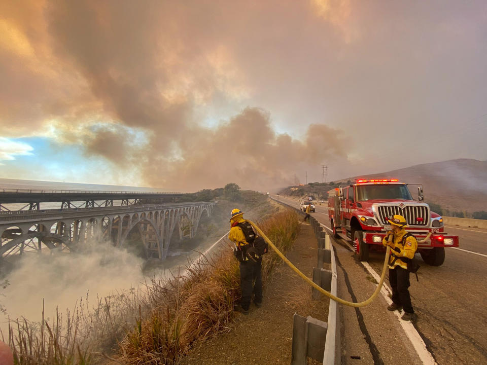 In this photo provided by the Santa Barbara County Fire Department, firefighters extinguish flames Tuesday afternoon, Oct. 12, 2021, that have spotted ahead of the fire front along Highway 101 southbound at Vista Point, north of Refugio State Beach in Santa Barbara County, Calif. (Mike Eliason/Santa Barbara County Fire Department via AP)