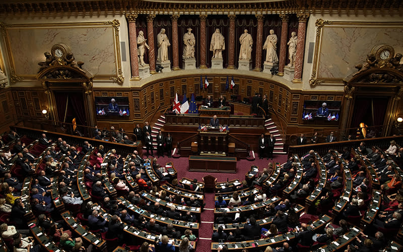 Britain's King Charles III addresses lawmakers from both the upper and the lower house of parliament in Paris. The photo is taken from a high angle, looking down at the whole room. King Charles III is standing at a podium in the center of the room, and before him is a semicircle of seats where the members of parliament sit.