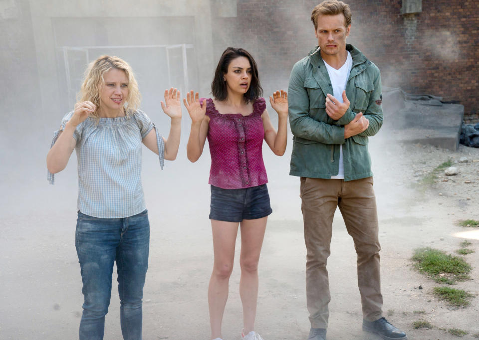 Kate McKinnon, Mila Kunis, and Sam Heughan holding their hands up in surrender in a scene from the movie