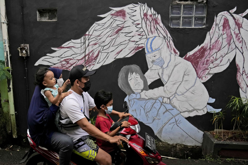 A family wearing masks to help curb the spread of coronavirus outbreak rides on a motorbike past a COVID-19-related mural honoring health workers in Jakarta, Indonesia, Saturday, Jan. 29, 2022. Indonesia is bracing for a third wave of COVID-19 infections as the highly transmissible omicron variant drives a surge in new cases, health authorities and experts said Saturday. (AP Photo/Dita Alangkara)