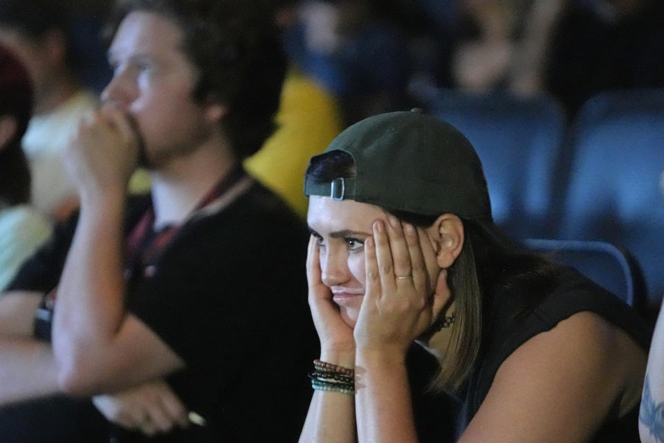 A person wearing a cap and bracelets, resting their face in their hands, appears deep in thought in an audience. Another person is nearby, also focused