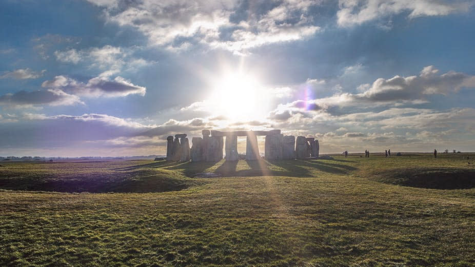  The sun shines through a blue cloudy sky over stonehenge surrounded by rolling grassy fields. 