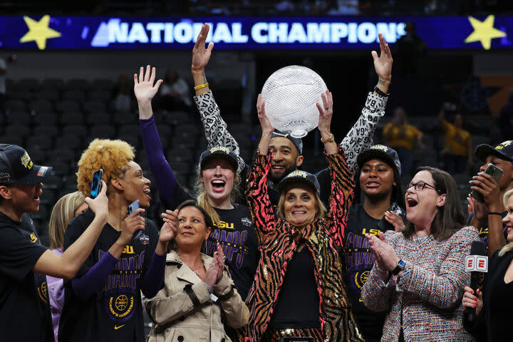 A group of women celebrate around a woman in the center of the photo raises a trophy above her head. A banner in the back reads "national championship."