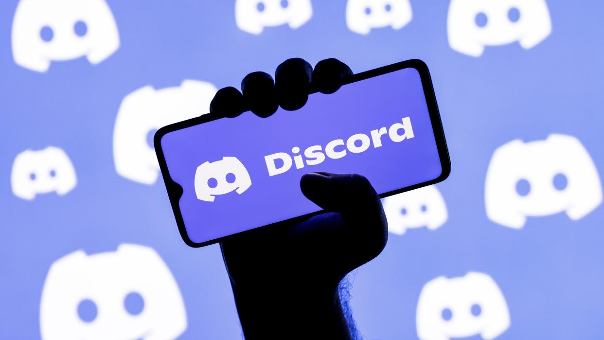  Smartphone with the Discord social gaming platform logo on the screen in a clenched hand on the background of Discord logos. 