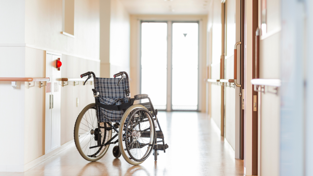 An empty wheelchair sits in the middle of a sunlit corridor of a nursing home.