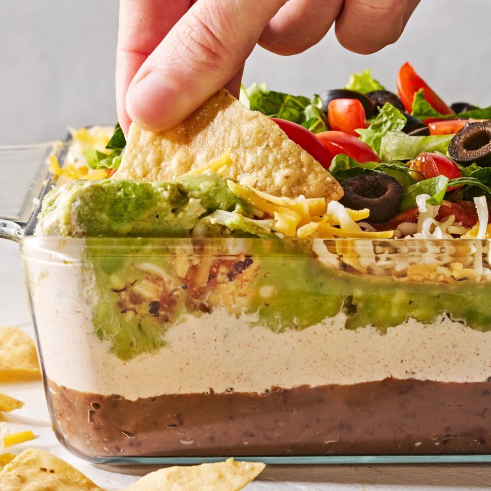 layered dip with refried beans, sour cream, guacamole, cheese, tomatoes black olives, and lettuce in a clear dish