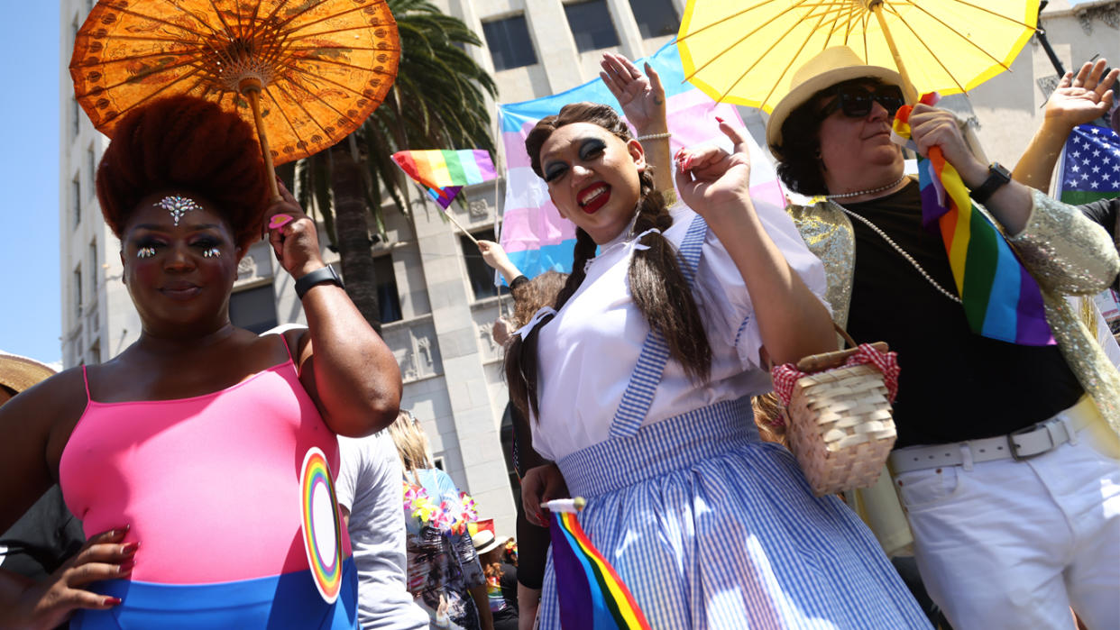 Twirling parasols and in fanciful makeup and costume, participants in the parade wave for the camera in front of an art deco building.