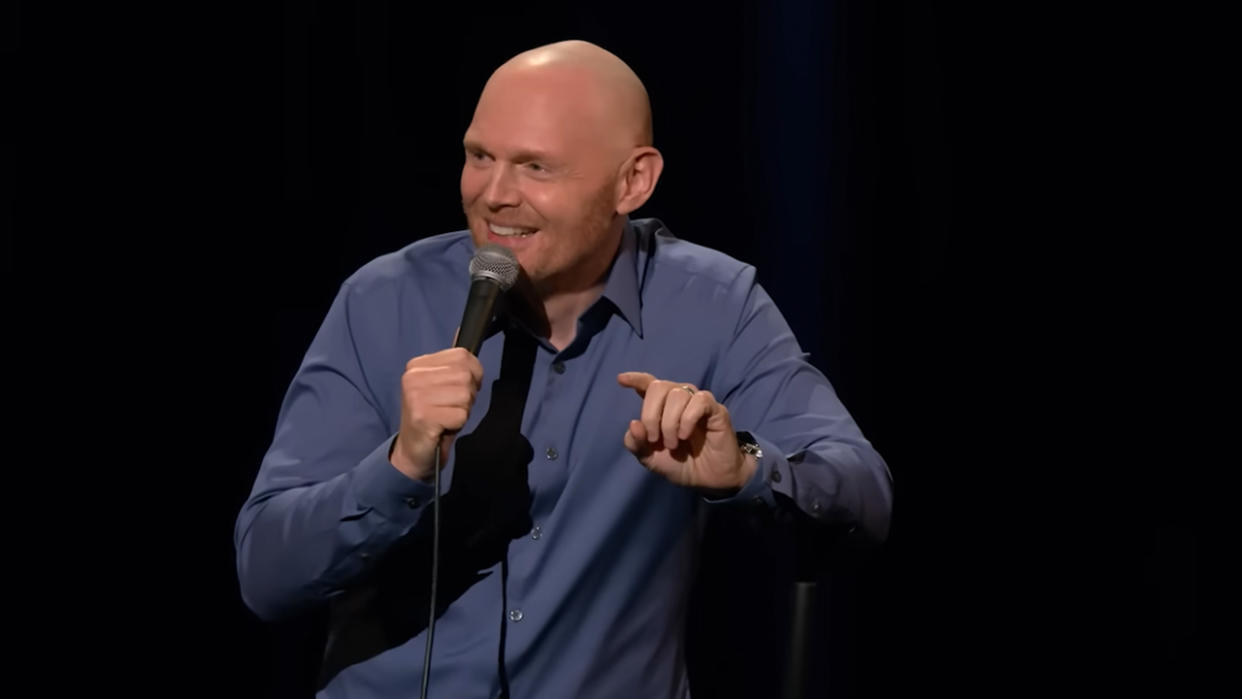  Bill Burr in a blue shirt doing standup, pointing and smiling. 