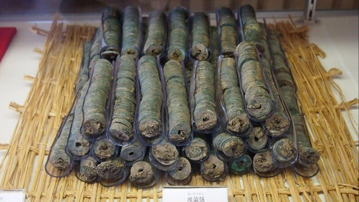  Photograph of stacks of bundled Japanese coins atop a straw woven mat upon a white tabletop. 