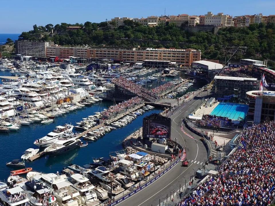 Französische Riviera voller Boote und Yachten neben der Grand-Prix-Rennstrecke mit einem roten Rennwagen, der gerade die Ziellinie überquert, neben Tribünen, die mit Hunderten von Fans gefüllt sind.