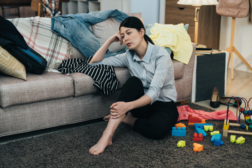 sleepy asian lady in blouse is sitting on floor with hand propping head on couch. japanese working mom taking rest in the parlor scattered with clothes and toys is feeling totally burnout.