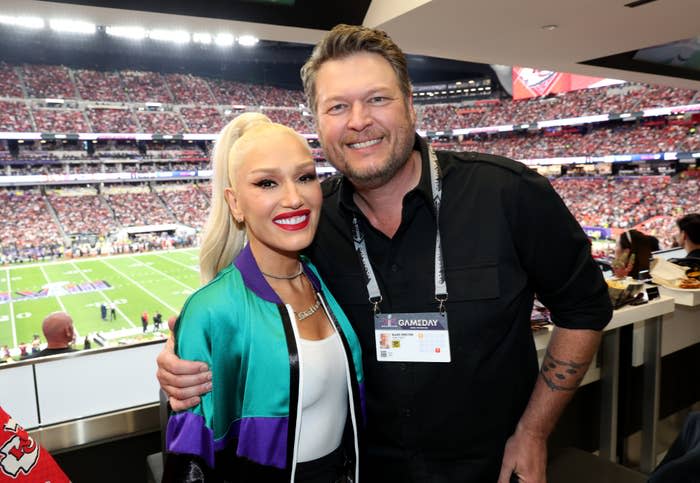 Gwen Stefani in a green and purple jacket smiling with Blake Shelton, in a black shirt, at a sports event