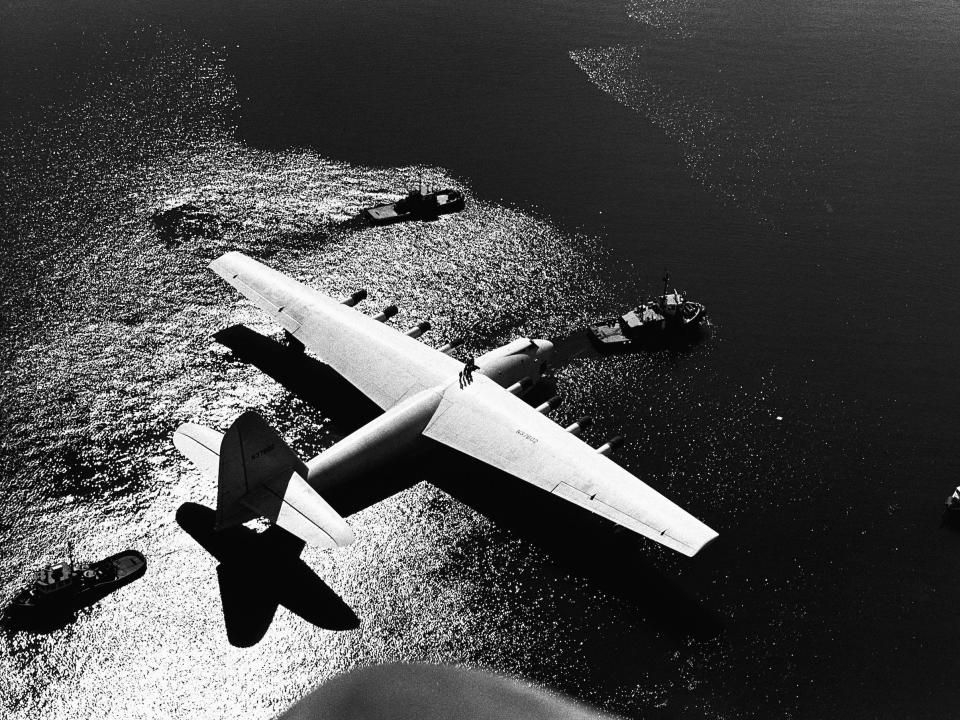 The Spruce Goose flying boat, designed by Howard Hughes, is towed to a temporary site at Long Beach before being exhibited to the public beside the Queen Mary liner.