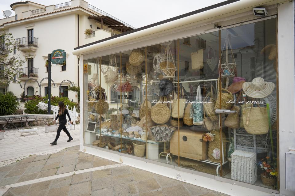 In this picture taken on Tuesday, April 28, 2020 a woman walks past a closed souvenir shop in the seaside town of Sperlonga, about 120km (80 miles) south of Rome. Normally, at this time of year Sperlonga would already be bustling with its first clients of the season. The restaurants would be fully operational and the golden-sand beaches, although still too chilly for Italians, would have been enjoyed by northern Europeans eager for some sunlight after a long, dark winter.(AP Photo/Andrew Medichini)