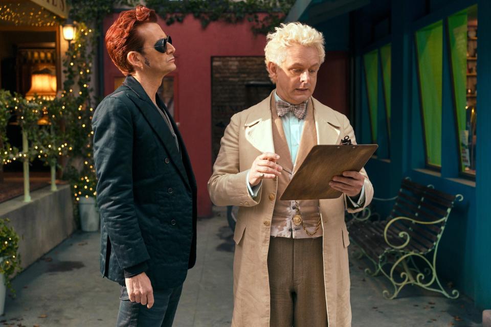 Two characters from "Good Omens," Crowley in aj acket and Aziraphale in a bow tie, examine a clipboard