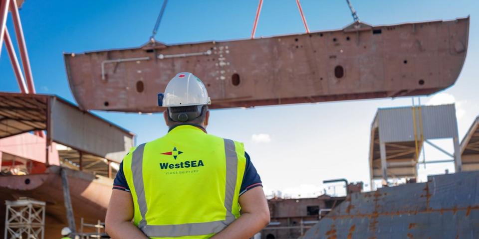 The keel being lifted in air by a crane with a WestSea shipyard person watching