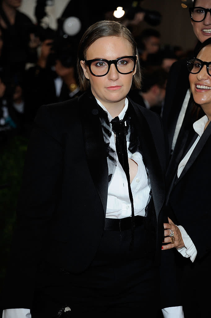 Lena Dunham wears a black tuxedo with a white shirt and black bow tie, standing on a red carpet at a celebrity event