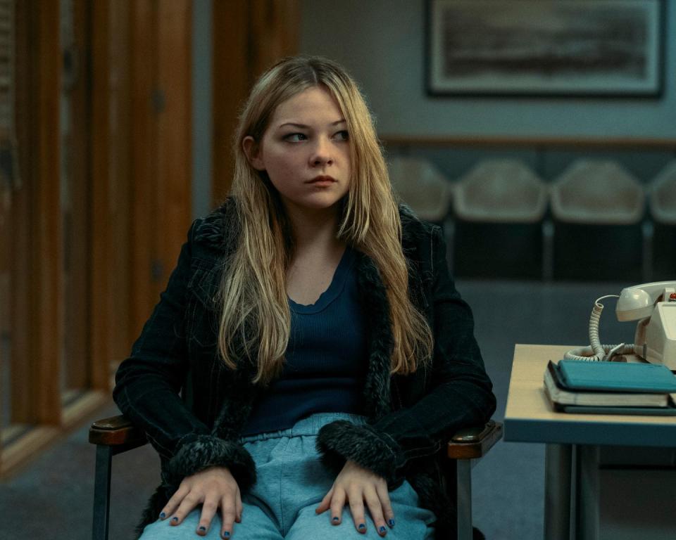 a girl sits in an armchair and looks to the right, she wears a sweater, a scoop neck shirt and pants, a desk with a phone and notebooks is on the right