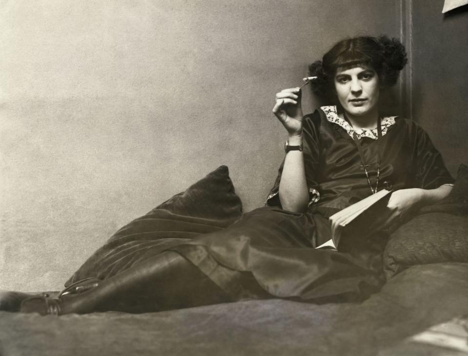 <p>A flapper woman relaxes on a chaise lounge with a cigarette in one hand and a book in the other. </p><p>"Flappers" were <a href="https://www.history.com/news/flappers-roaring-20s-women-empowerment" rel="nofollow noopener" target="_blank" data-ylk="slk:typically young women;elm:context_link;itc:0;sec:content-canvas" class="link ">typically young women</a> in the 1920s who enjoyed jazz and cigarettes while sporting a "bob" hairstyle and a knee-length skirt.</p>