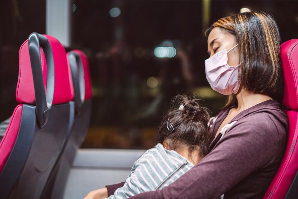 Young mom in protective face mask holding her sleeping daughter in her arms while riding on bus.