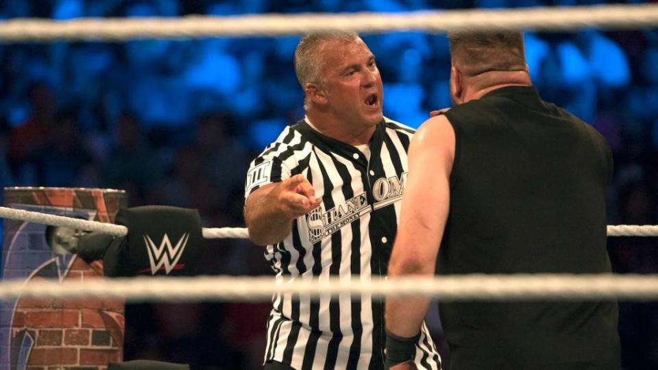 An angry-looking middle-aged man in a referee shirt yells at another wrestler, gesturing to him to step back. They are in a wrestling ring in front of a large crowd.