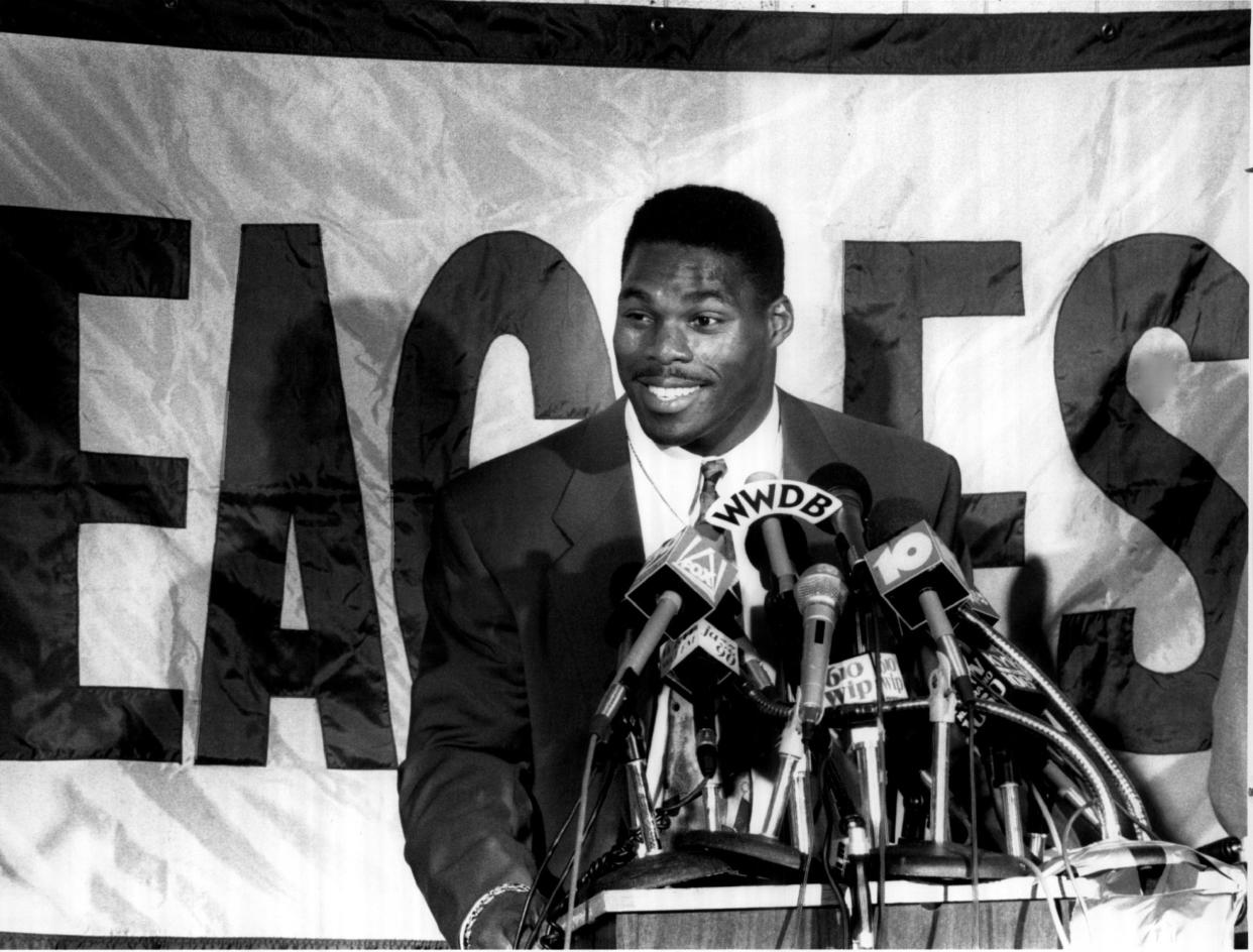 Herschel Walker talks in front of a bank of microphones, with a banner saying Eagles behind him.