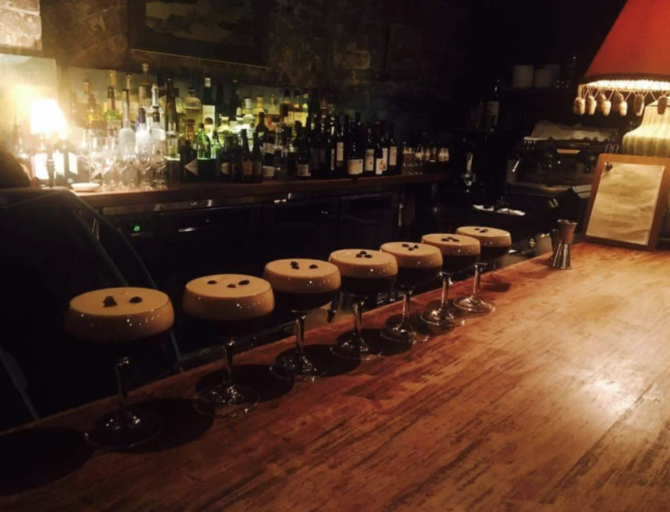 Dimly lit bar with stools lined up, empty wine glasses on the counter, and shelves of bottles in the background