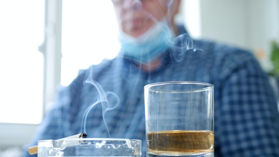 un verre court contenant de l’alcool à côté d’un cendrier dans lequel se trouve une cigarette fumante. en arrière-plan, hors champ, un homme portant une chemise bleue et un masque chirurgical