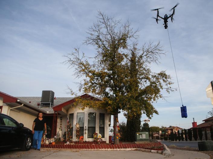 A woman in a black top and blue jeans waits for a drone to drop a package in the driveway of her small home in Texas.
