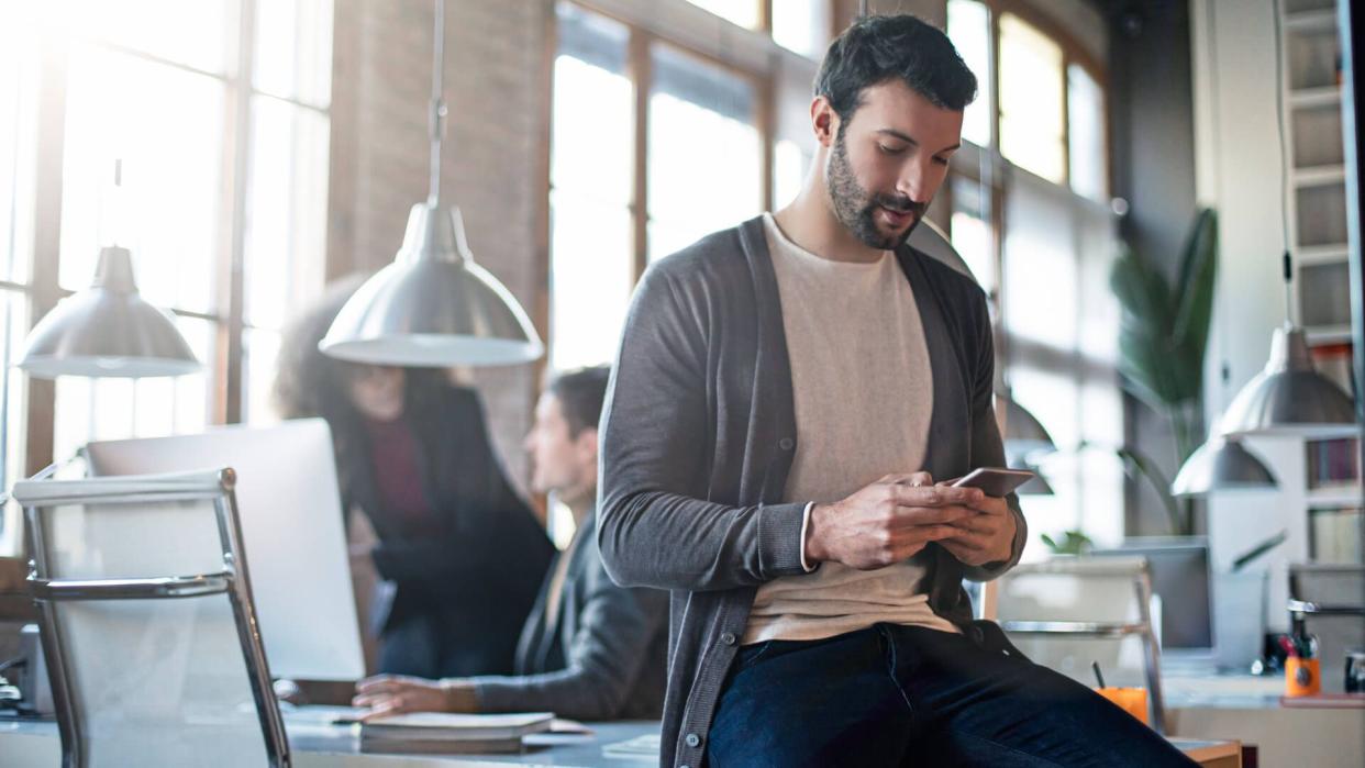 Businessman checking the mobile phone sitting on his desk, colleagues working in the background.