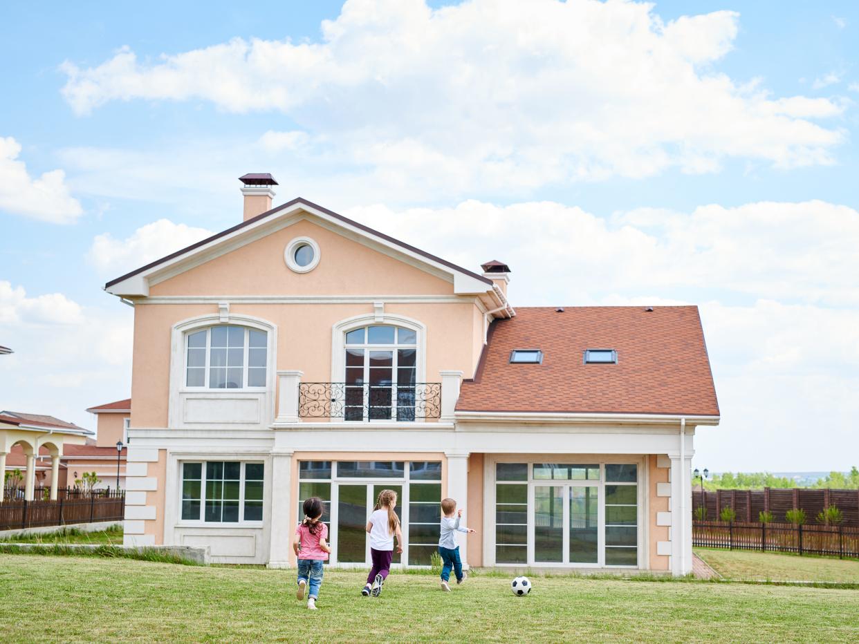 Wide angle portrait of three happy little kids playing football in front yard of beautiful two storey house